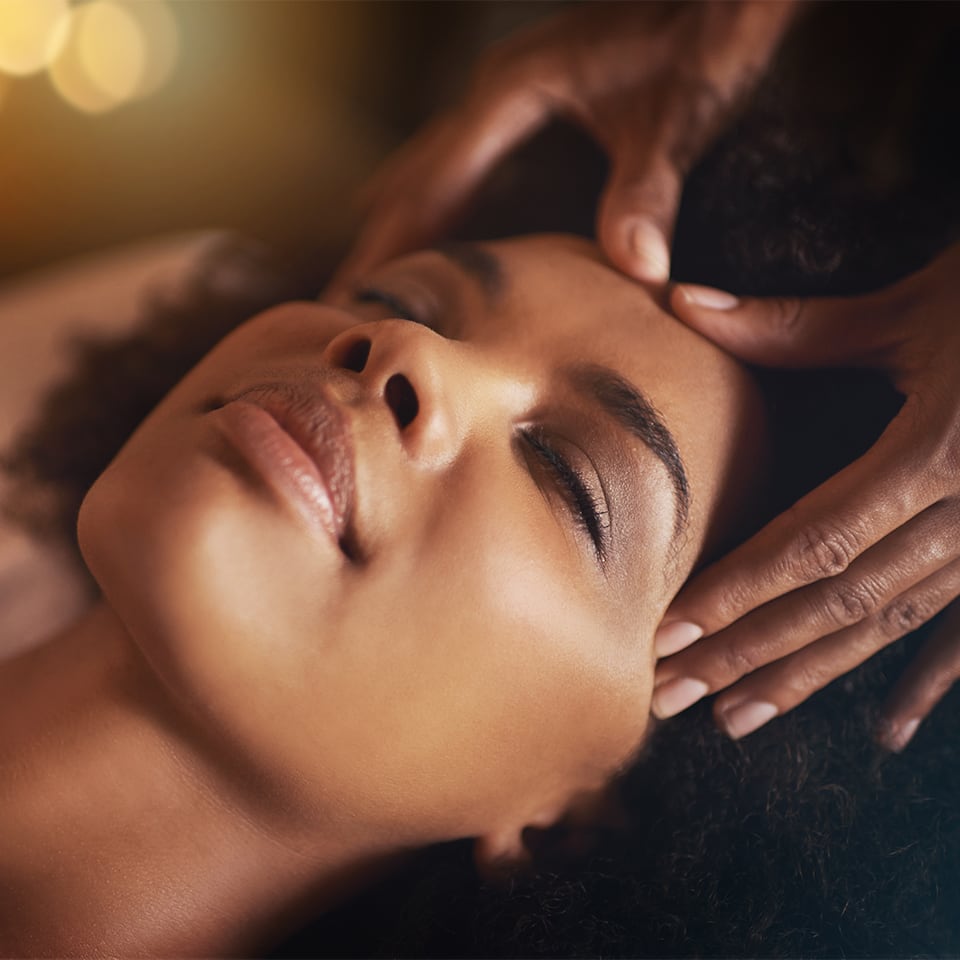 Closeup of a Black woman’s face as she receives a massage to the temples and forehead; her eyes are closed and she is deeply relaxed